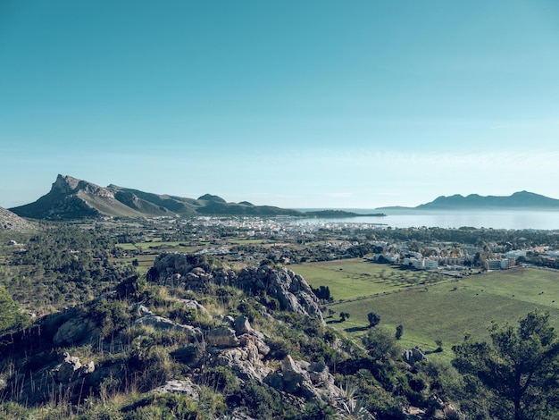 Pittoresca vista drone della città di Pollensa situata tra verdi campi agricoli e montagne contro il cielo blu sulla costa del mare a Maiorca, Spagna