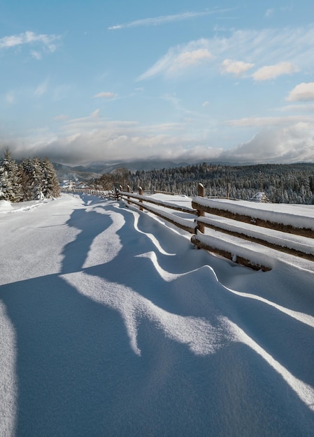 Pittoresche ombre ondulate sulla neve da una recinzione di legno Alpine montagna villaggio invernale periferia sentiero innevato foresta di abeti Immagine ad alta risoluzione con grande profondità di campo