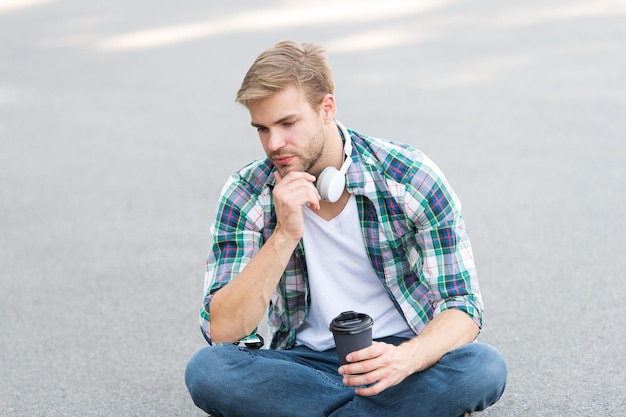 Più caffè. caffè da portar via. ragazzo beve caffè all'aperto. l'uomo si siede a terra. studente stanco in cuffia. formazione in linea. ascoltare musica. concetto di e-book. camicia a scacchi uomo. studente rilassarsi e ricaricarsi.