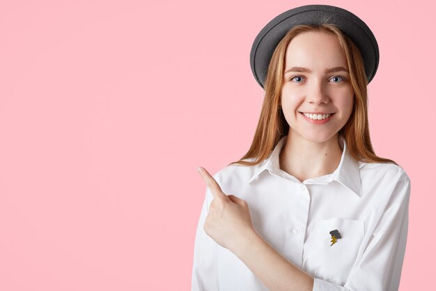 Piuttosto felice giovane femmina dagli occhi blu con i capelli lunghi, gli occhi azzurri, indossa un elegante cappello nero e camicia bianca, indica lo spazio vuoto sulla parete rosa per la tua pubblicità o testo promozionale