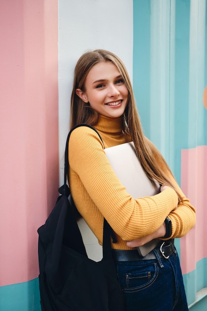 Piuttosto gioiosa studentessa casual ragazza con il computer portatile felicemente guardando a porte chiuse su sfondo colorato all'aperto