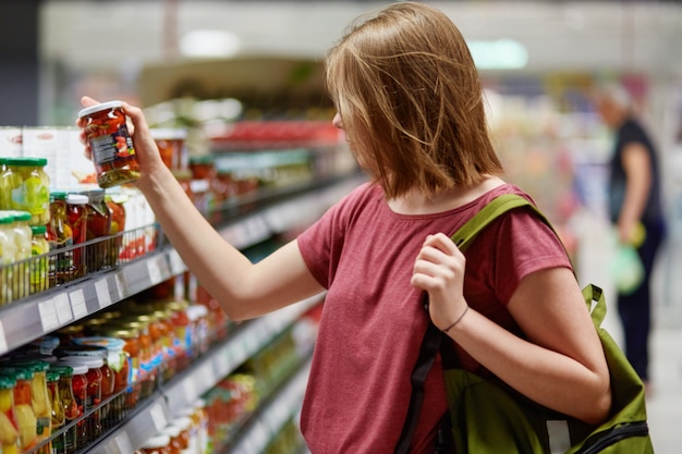 piuttosto giovane cliente femake detiene merci in scatola in un contenitore di vetro, si trova in un negozio di alimentari, vestito con abiti casual