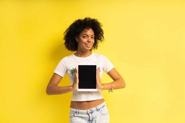 Piuttosto giovane donna afro-americana in piedi e utilizzando computer tablet isolate su sfondo giallo.