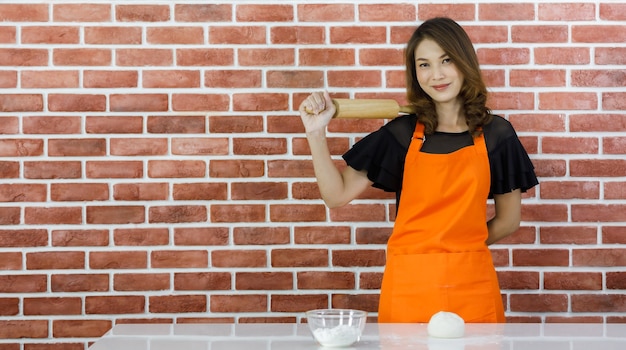 Piuttosto giovane donna asiatica in grembiule arancione sorridente in piedi vicino al muro di mattoni e posa divertente scherzando mostrando mattarello di legno nella cucina di casa
