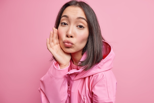 Piuttosto giovane donna asiatica tocca la guancia tiene le labbra piegate vuole baciarti ha un aspetto tenero pelle sana capelli scuri indossa una giacca elegante con cappuccio isolato sul muro rosa. Espressioni facciali