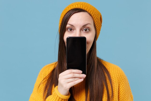 Piuttosto giovane donna bruna ragazza in maglione giallo e cappello in posa isolata su sfondo blu. Concetto di stile di vita delle persone. Mock up spazio di copia. Faccia coprente con il telefono cellulare con schermo vuoto vuoto.