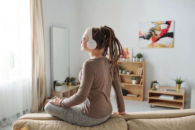 Piuttosto giovane donna casual con i dreadlocks godendo la musica di meditazione in cuffie mentre era seduto sul divano