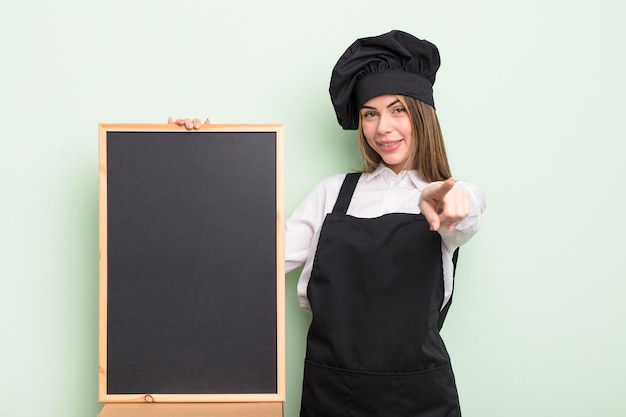 Piuttosto giovane donna che punta alla telecamera scegliendo te. chef con lavagna