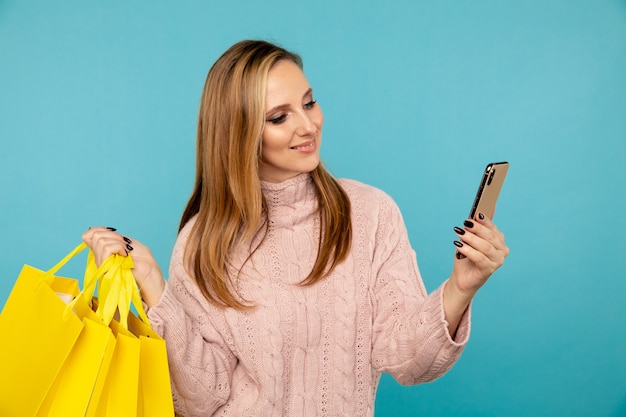 Piuttosto giovane donna con borse gialle e telefono isolato su sfondo blu. Concetto di vendita.