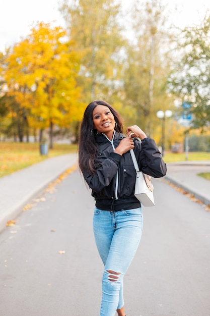 Piuttosto giovane donna di colore felice in una giacca casual alla moda e jeans blu con una borsetta cammina nel parco con fogliame autunnale giallo colorato