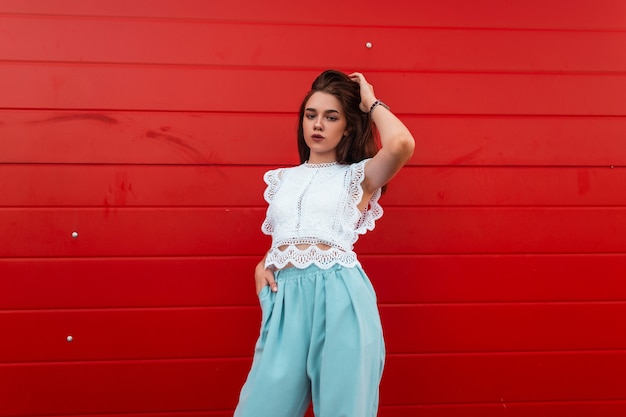 Piuttosto giovane donna europea in un'elegante camicia di pizzo in pantaloni blu con capelli castani si trova in città vicino a un muro di legno rosso vintage.