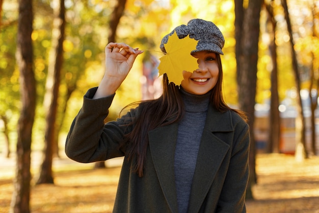 Piuttosto giovane donna felice alla moda con un sorriso in un cappotto alla moda con un cappello vintage con una foglia d'autunno gialla copre il viso nel parco