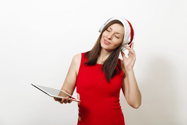 Piuttosto giovane donna felice in abito rosso e cappello di Natale che ascolta musica in cuffia, tenendo tablet su sfondo bianco. Aggeggio della ragazza di Santa isolato. Vacanze di Capodanno 2018. Annuncio dello spazio di copia.
