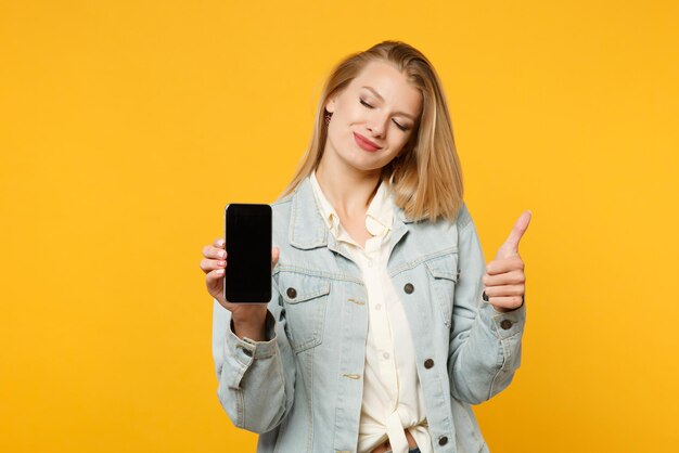 Piuttosto giovane donna in abiti casual denim che mostra il pollice in su, tenendo il telefono cellulare con schermo vuoto vuoto isolato su sfondo giallo arancione in studio. Concetto di stile di vita delle persone. Mock up spazio di copia.