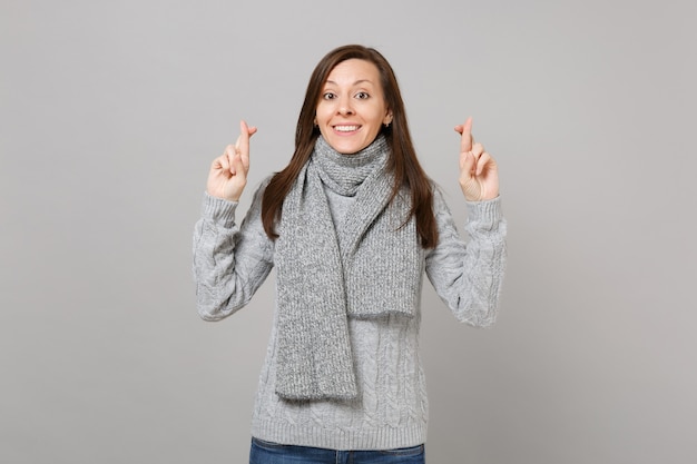 Piuttosto giovane donna in maglione grigio, sciarpa tenendo le dita incrociate, esprimendo desiderio isolato su sfondo grigio. Stile di vita sano, persone sincere emozioni, concetto di stagione fredda. Mock up copia spazio.