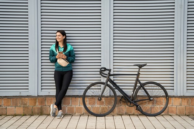Piuttosto giovane donna in piedi vicino al muro grigio con la bici e tenendo lo smartphone all'aperto. Concetto di sport e stile di vita