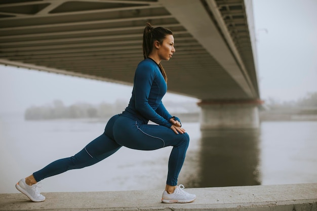 Piuttosto giovane donna in tuta blu che si estende prima dell'allenamento in riva al fiume al mattino d'autunno