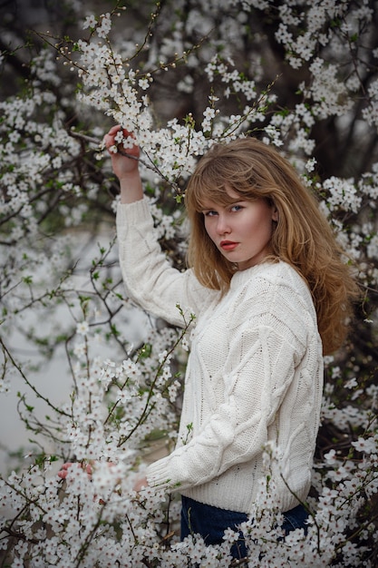 Piuttosto giovane donna in un maglione bianco nel giardino di fiori di ciliegio, tempo di primavera