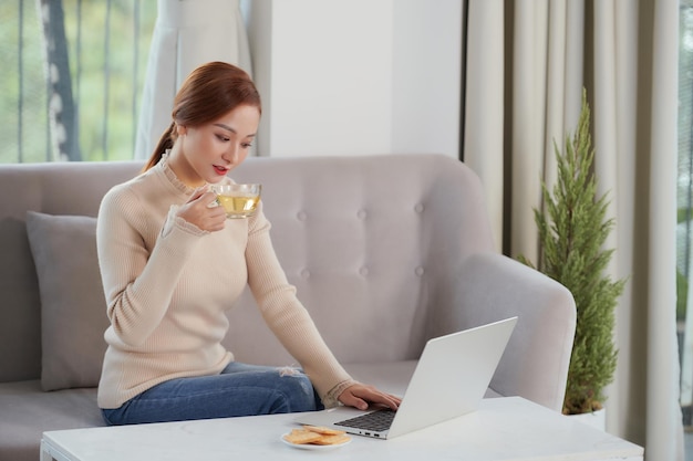 Piuttosto giovane donna vietnamita che beve una tazza di tè e lavora al computer portatile quando si siede sul divano a casa