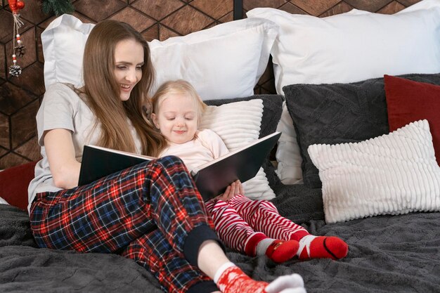 Piuttosto giovane madre che legge un libro a sua figlia a letto la vigilia di Natale Mamma o baby sitter che raccontano fiabe a una bambina in età prescolare