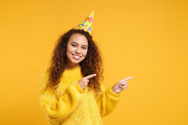 Piuttosto giovane ragazza afroamericana in maglione di pelliccia, cappello di compleanno in posa isolato su sfondo giallo arancione ritratto in studio. Concetto di stile di vita delle persone. Mock up spazio di copia. Puntando gli indici da parte.