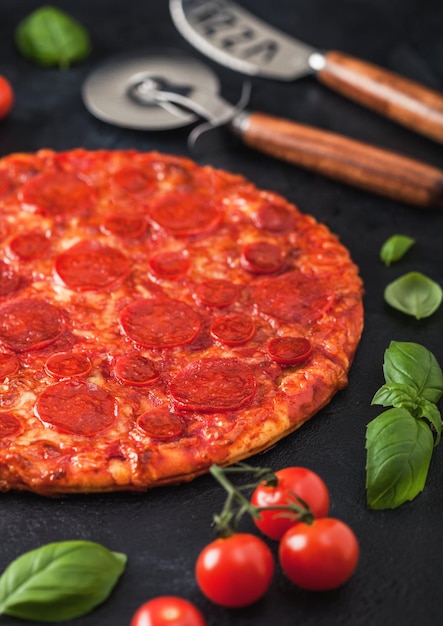 Pizza ai peperoni calda e piccante fresca al forno con taglierina e coltello con pomodori e basilico su sfondo nero del tavolo da cucina