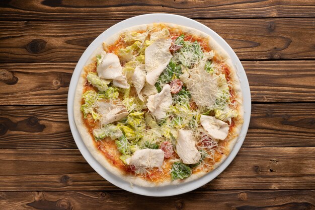 Pizza Caesar su pasta sottile con petto di pollo, lattuga, pomodorini e parmigiano in un piatto di ceramica bianca su un tavolo da cucina in legno.