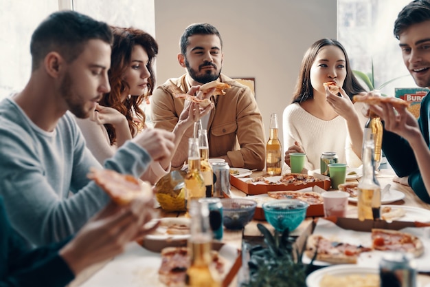 Pizza deliziosa. Gruppo di giovani in abbigliamento casual che mangiano pizza mentre fanno una cena al chiuso