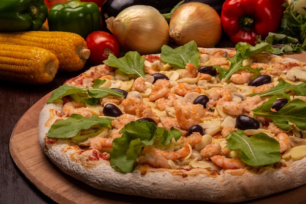 Pizza di gamberi e funghi con rucola su una tavola di legno e verdure in sottofondo.