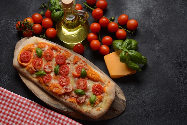 Pizza focaccia guarnita con pomodorini freschi, basilico, formaggio e olio d'oliva su tavola per pizza in legno. Sfondo scuro