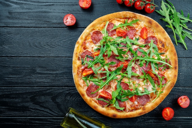 Pizza fresca fatta in casa con salame, pomodorini e rucola su un legno nero con spazio di copia.