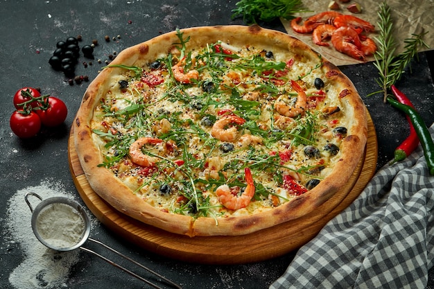 Pizza italiana cotta al forno con salsa, formaggio, rucola, gamberetti e olive in una composizione con ingredienti su un tavolo scuro. Vista dall'alto Pizza con frutti di mare