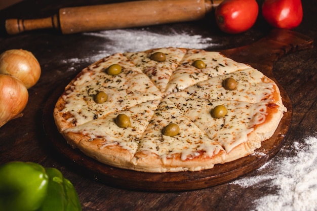 Pizza mozzarella con olive con diverse verdure intorno su una tavola di legno.
