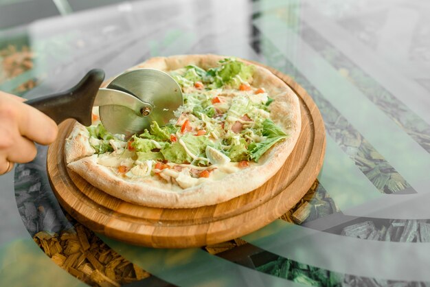 Pizza per affettare utilizzando il coltello a ruota pizza con prosciutto, salame, pomodori, insalata e parmigiano su una tavola di legno su un tavolo di vetro