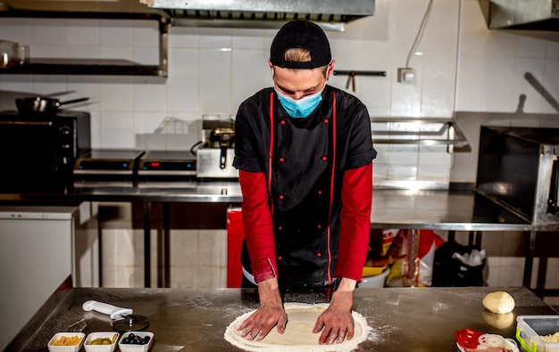 Pizzaiolo o produttore con una maschera medica contro il coronavirus, realizza degli spazi vuoti per la pasta della pizza Impasto per pizza rendendo il concetto