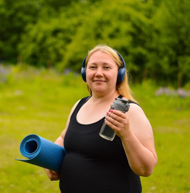 Plus size donna caucasica bianca in cuffia una maglietta nera con un tappetino da yoga in mano sta camminando per strada