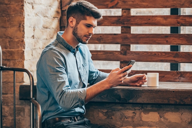 Pochi minuti per un caffè e un messaggio veloce. Vista laterale del bel giovane premuroso che tiene in mano lo smartphone e lo guarda mentre è seduto vicino alla finestra nell'interno del loft con una tazza di caffè in mano