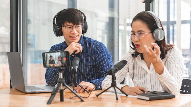 Podcaster asiatico uomo e donna in cuffie che registrano contenuti con un collega che parla al microfono e fotocamera in studio di trasmissione insieme, tecnologia di comunicazione e concetto di intrattenimento