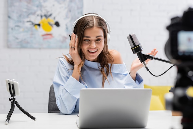 Podcaster bruna spensierata con le cuffie che parla e guarda il portatile vicino al microfono e sfocata