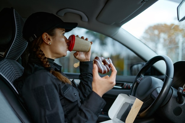 Poliziotto femminile che pranza durante la giornata lavorativa. Ufficiale di polizia della donna che beve caffè, mangiando la ciambella in macchina