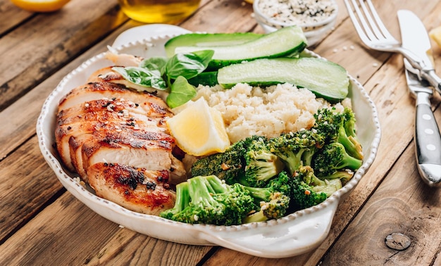 Pollo alla griglia con broccoli di riso al cavolfiore e cetriolo in una ciotola bianca su un tavolo di legno rustico Ciotola per il pranzo Keto Vista dall'alto