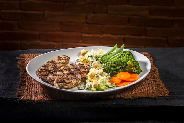 Pollo alla griglia, insalata caesar e verdure su panno marrone e legno nero su sfondo di mattoni.