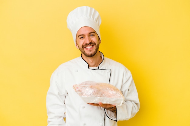 Pollo della holding dell'uomo giovane chef caucasico isolato su sfondo giallo felice, sorridente e allegro.