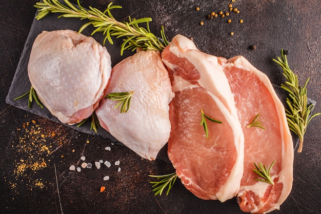 Pollo e maiale crudi su ardesia con spezie. Sfondo scuro, vista dall&#39;alto, copia spazio.