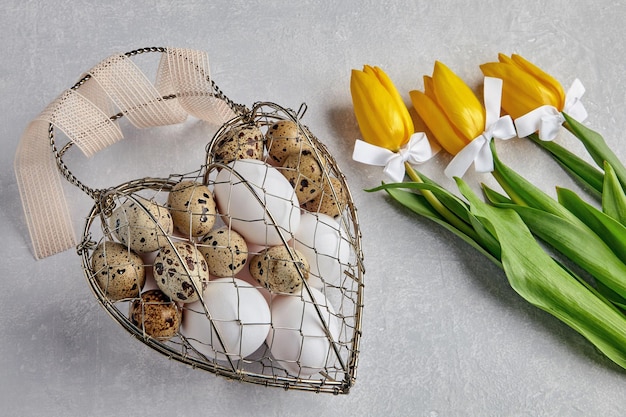 Pollo e uova di quaglia in una scatola di metallo di vimini a forma di cuore legata con un nastro e tulipani gialli su uno sfondo di cemento chiaro