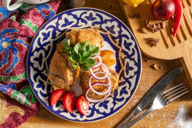 Pollo giovane alle spezie, cotto fino a doratura con patate, cipolle rosse e pomodori in un piatto con ornamenti tradizionali uzbeki