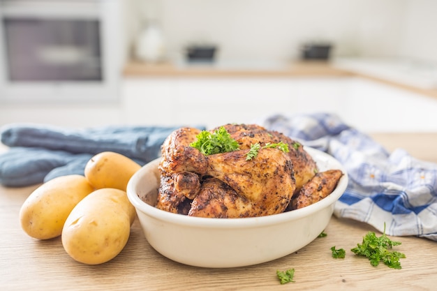 Pollo intero arrosto in un piatto bianco con patate crude sul tavolo cucinato. Interno di casa in background.