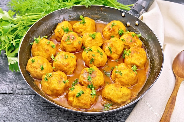 Polpette con salsa di pomodoro in una padella, prezzemolo, aneto, tovagliolo e cucchiaio su uno sfondo di tavola di legno