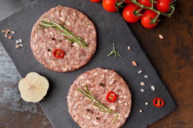 Polpette di hamburger di manzo macinato fresco crudo, verdure al rosmarino e spezie su un piatto di pietra nera su sfondo scuro vista dall'alto Prodotti semilavorati di carne per un hamburger fatto in casa