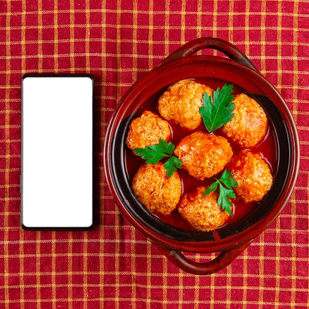 Polpette servite con salsa di pomodoro in pentola di coccio e smartphone con schermo bianco vuoto